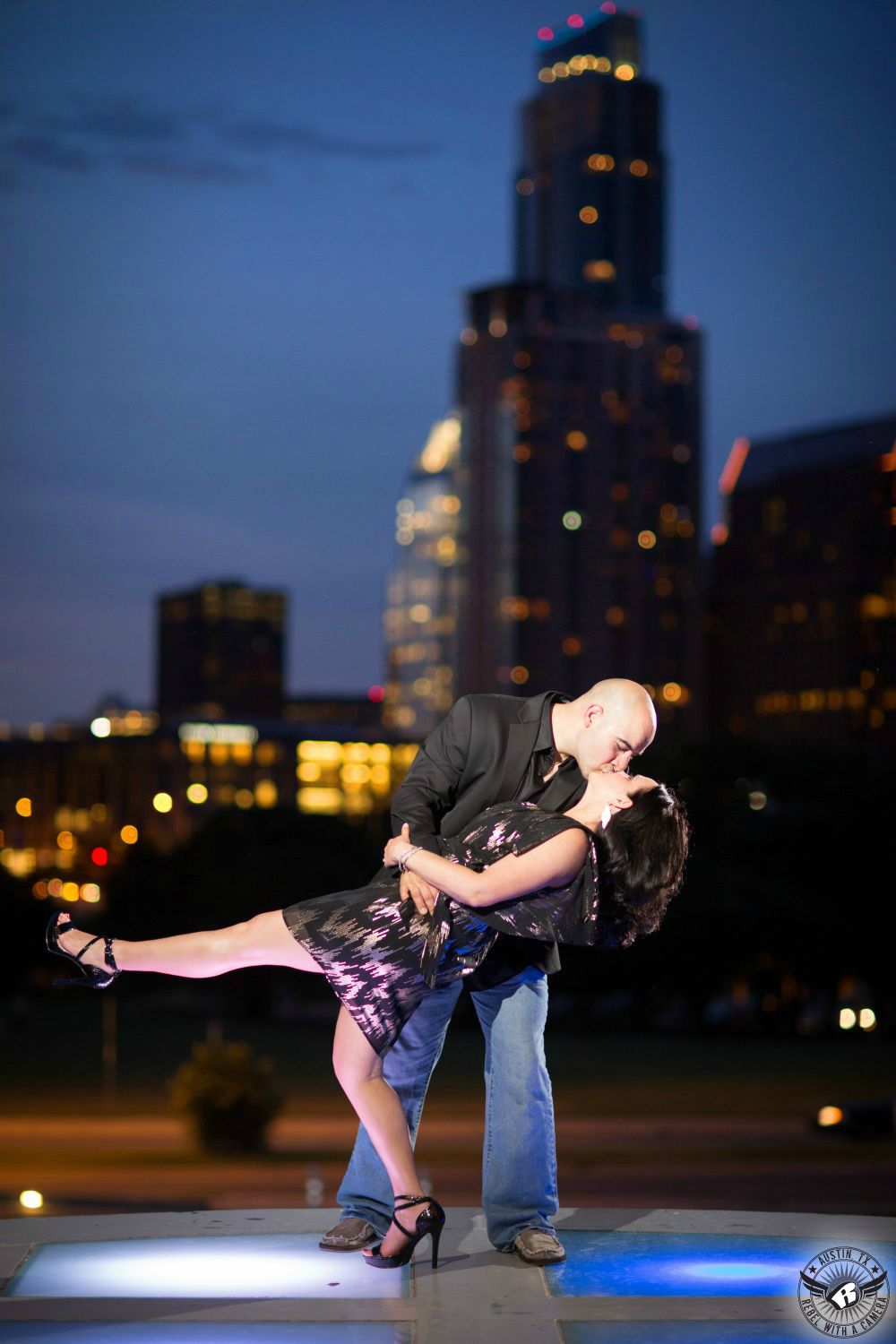 latino guy with shaved head and black shirt and blue jeans dips brunette girl in purple and black dress and black high heals on a lit up floor in front of the skyline of austin at night with a blue and purple sky in this engagement portrait 
